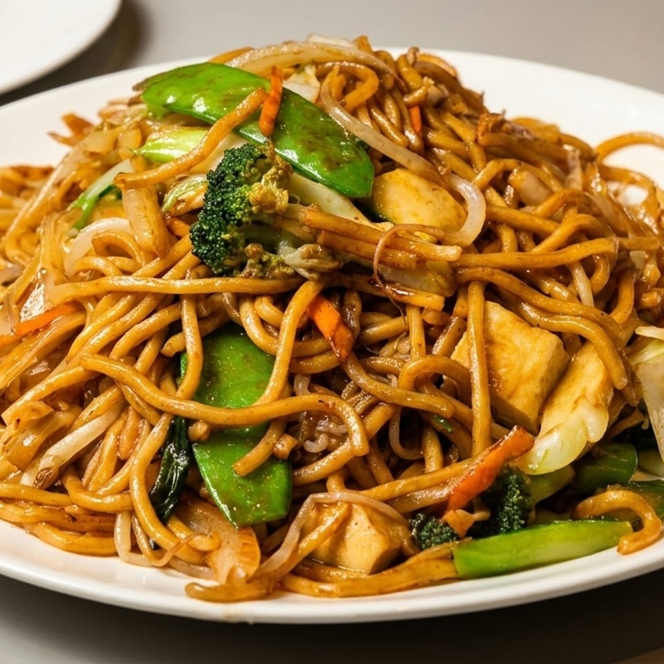 A white plate filled with vegetable lo mein noodles, including broccoli, snap peas, and carrots.