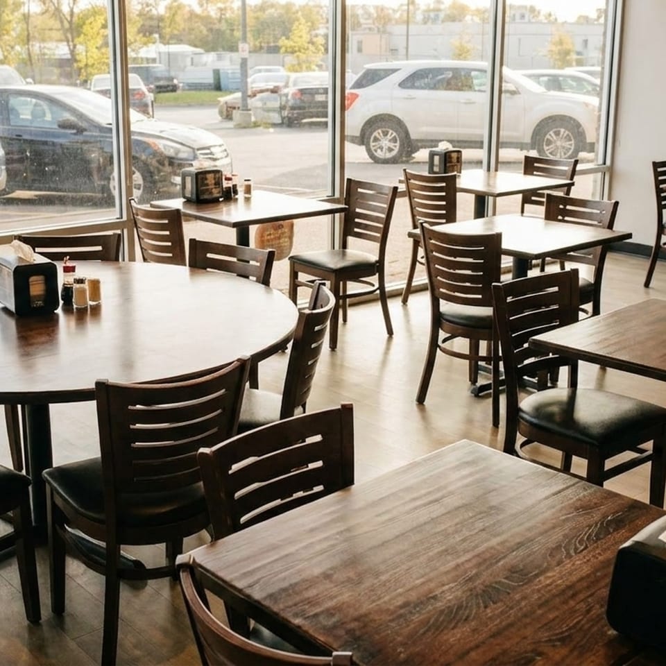 Empty restaurant dining room with dark wooden tables and chairs, and large windows overlooking a parking lot.